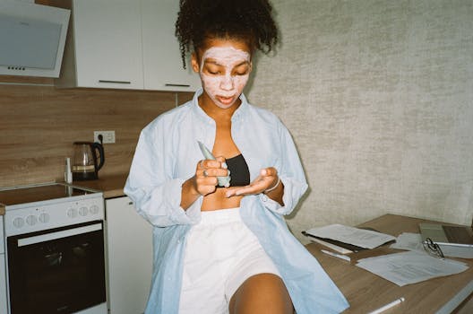 A woman applying facial cream sits on a kitchen counter while working remotely on a laptop.