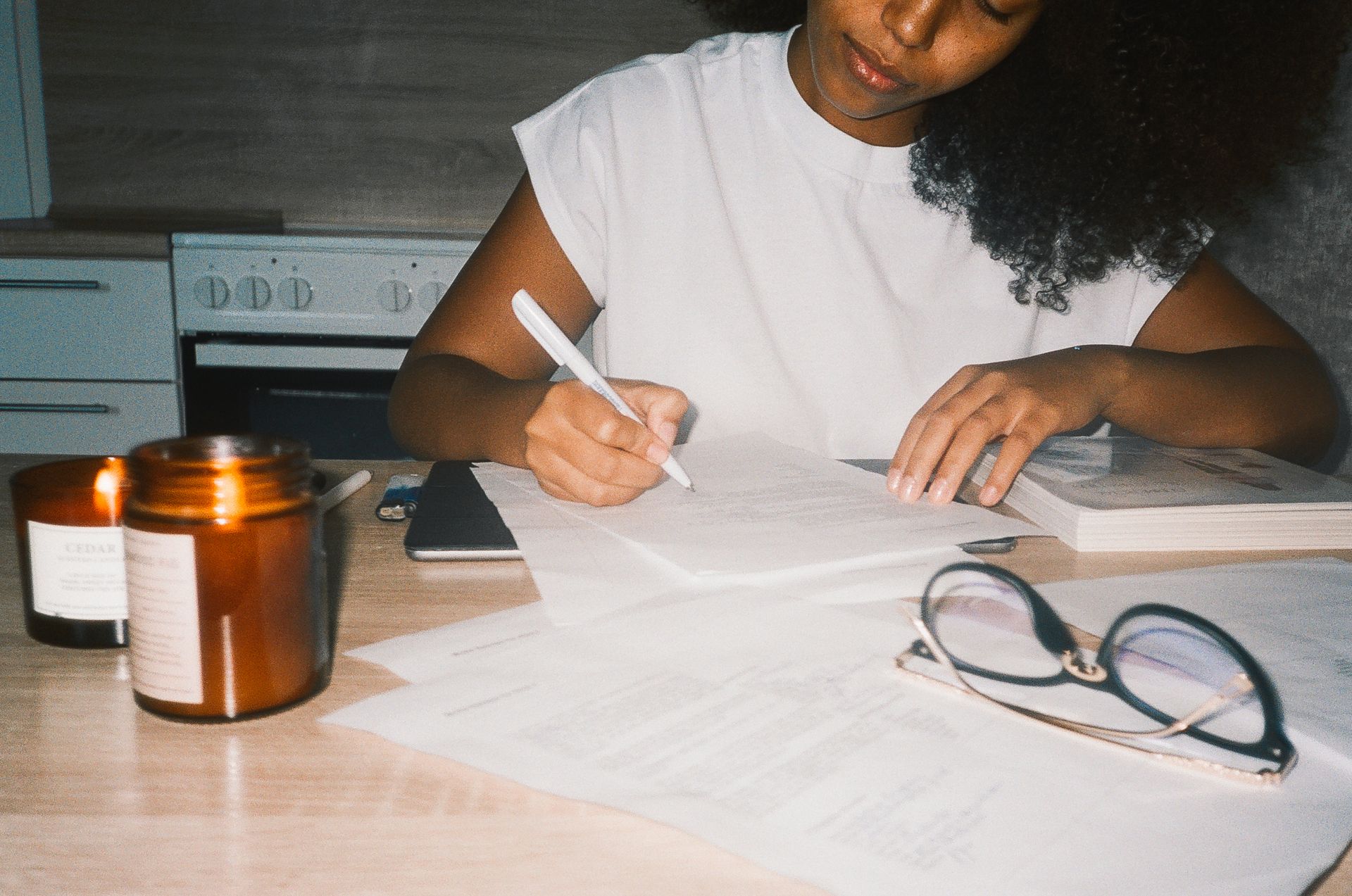 A black woman focused on remote work, writing notes by candlelight at home.