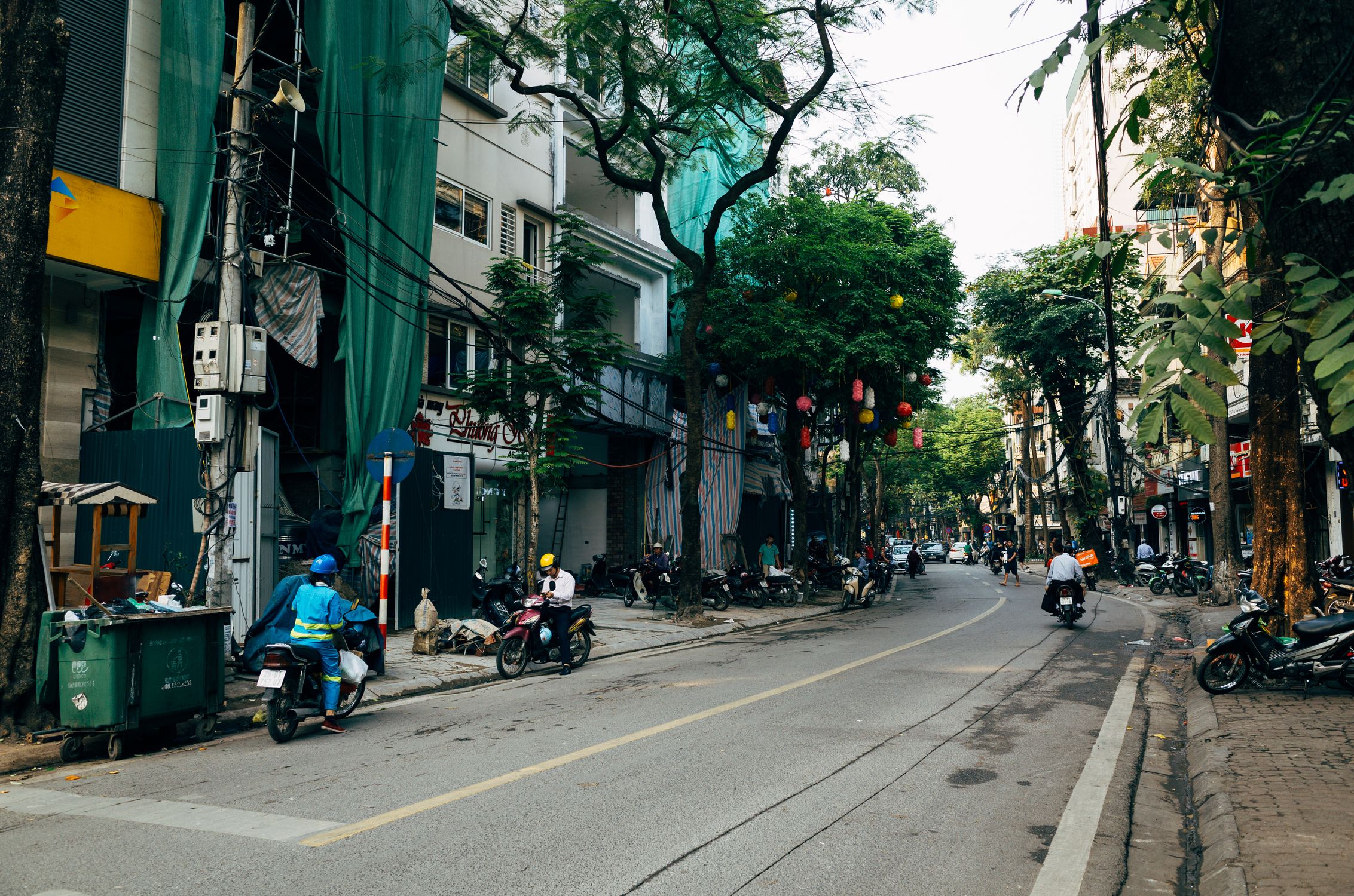 Vibrant Hanoi street scene with motorbikes, trees, and urban life in Vietnam.