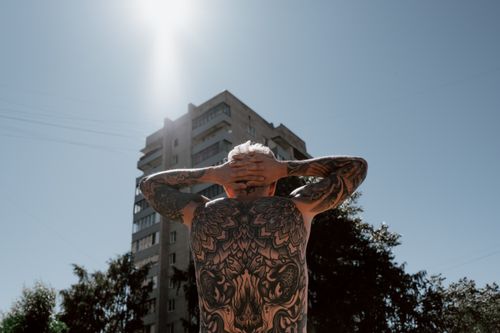 Free Back view of a tattooed person standing outdoors near an urban building under clear skies. Stock Photo