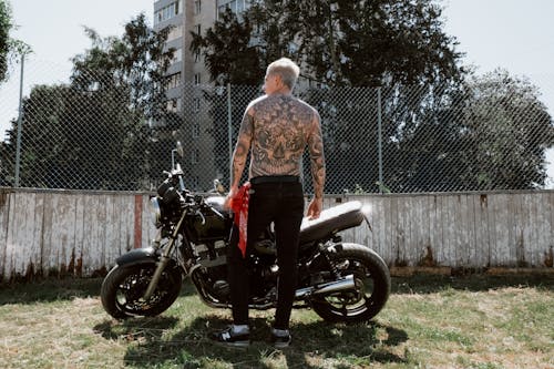 Free Shirtless tattooed man standing beside motorcycle in urban outdoor setting, exuding adventure. Stock Photo