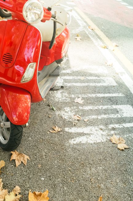 Red scooter parked on dry autumn pavement with scattered leaves, suggesting a peaceful fall day.