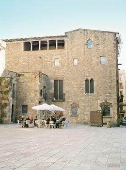 Café patrons enjoy a sunny afternoon by a historic stone building, creating a cozy urban setting.