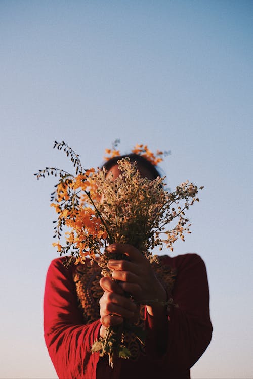 Unrecognizable female in bright apparel covering face with dry autumn bouquet under blue sky