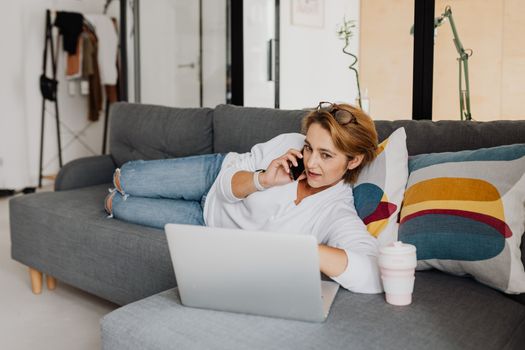 Woman lying on a couch using her phone and laptop, working remotely from home.
