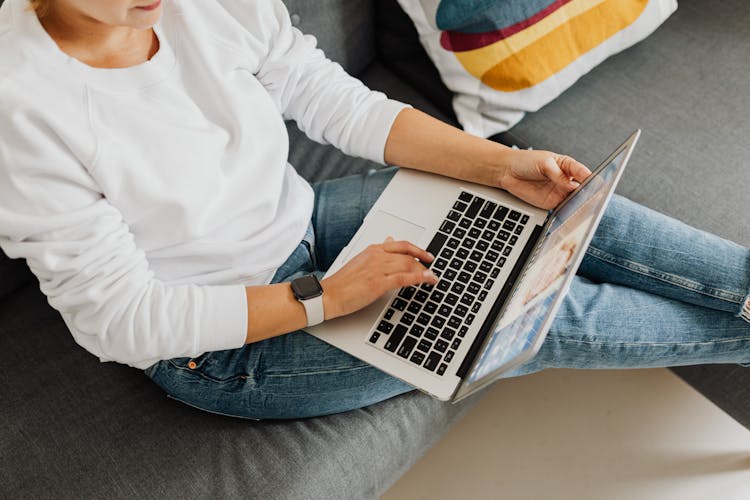 Person In White Sweater Sitting On Gray Couch Using Laptop