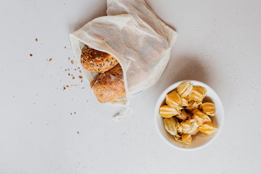 A flat lay of bread in a reusable bag and fruits in a bowl on a white surface, promoting zero waste.