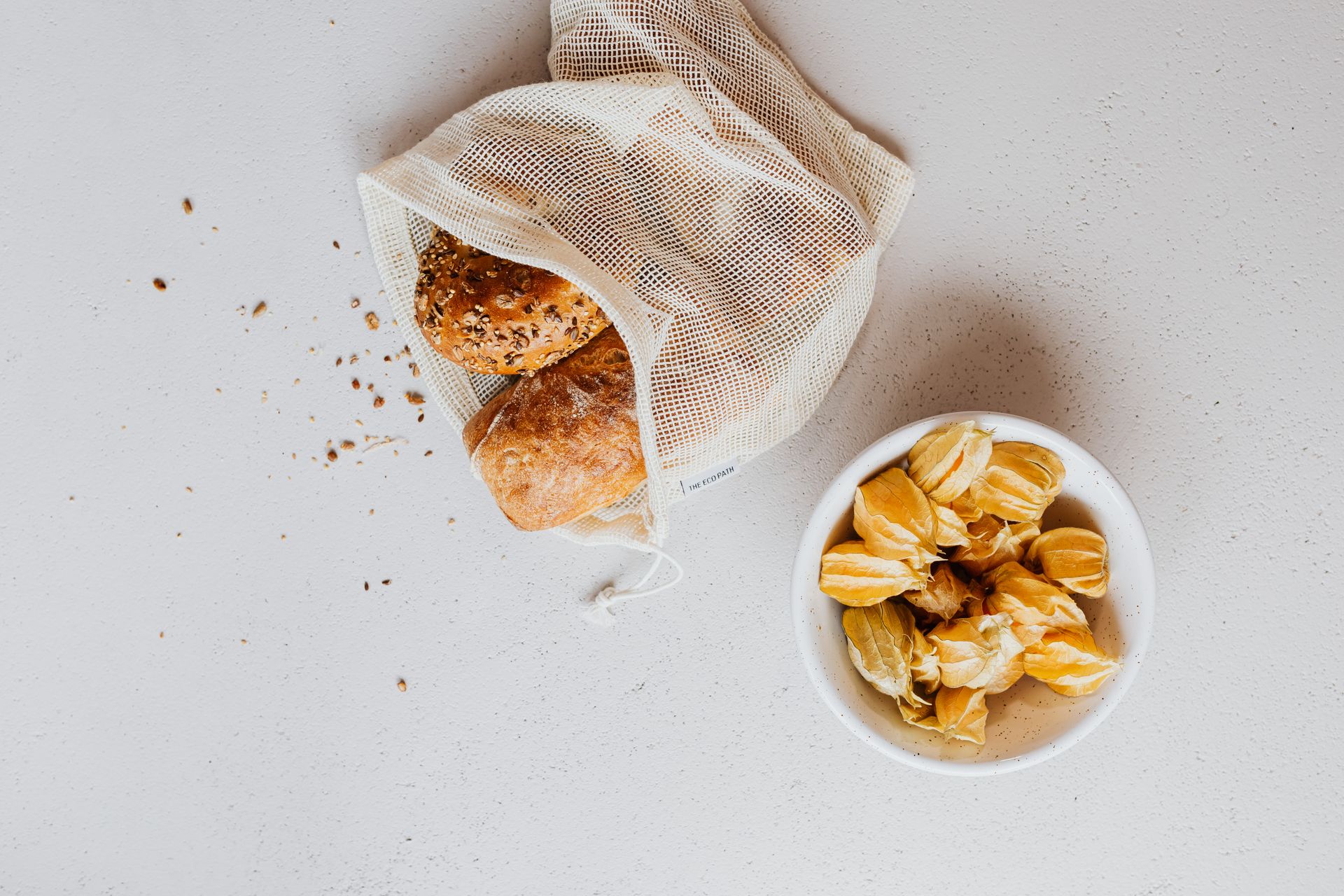 A flat lay of bread in a reusable bag and fruits in a bowl on a white surface, promoting zero waste.