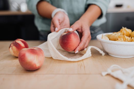 Hands arranging fresh peaches in a reusable net bag on a wooden table for zero waste.