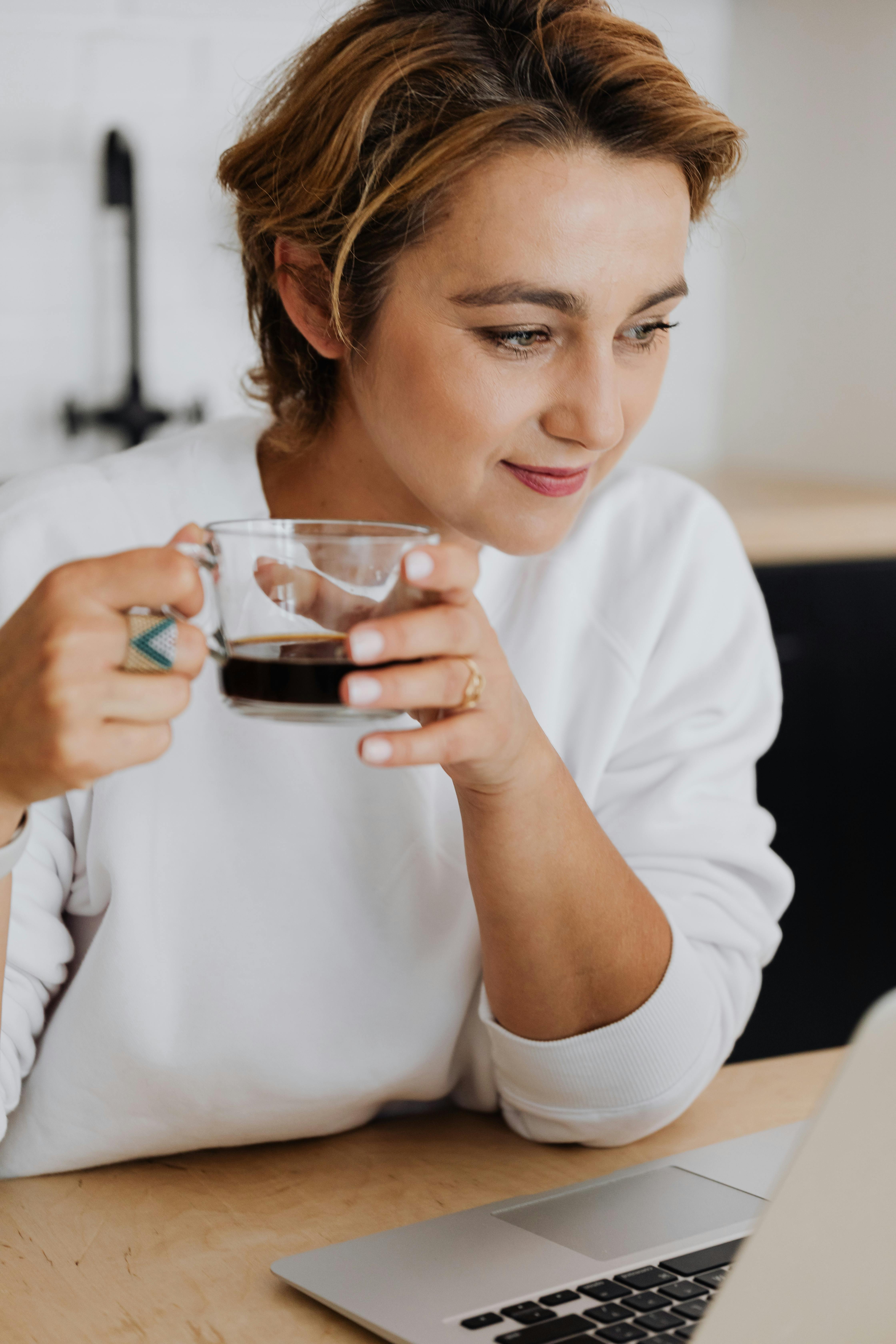 Focused woman holding coffee and using a laptop, wearing white in a cozy setting.