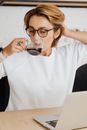 一位戴着眼镜的女人在室内喝着咖啡，用笔记本电脑工作
