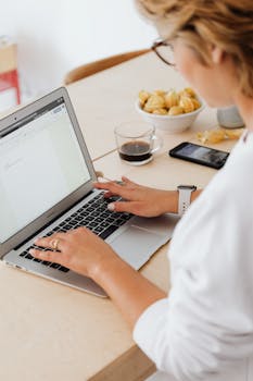 Adult woman working from home, typing on laptop with coffee and snacks on wooden table.