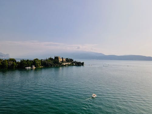 From above of rippled sea with motorboat near old building with trees and ridge silhouettes under cloudy sky