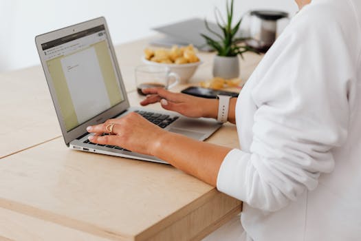 Close-up of a woman typing on a laptop at a home office desk with coffee and snacks.
