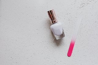 Flat lay of white nail polish and pink tinted glass nail file on a white textured background, ideal for beauty themes.
