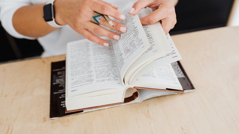 A close-up of a woman turning the pages of a dictionary on a wooden desk.
