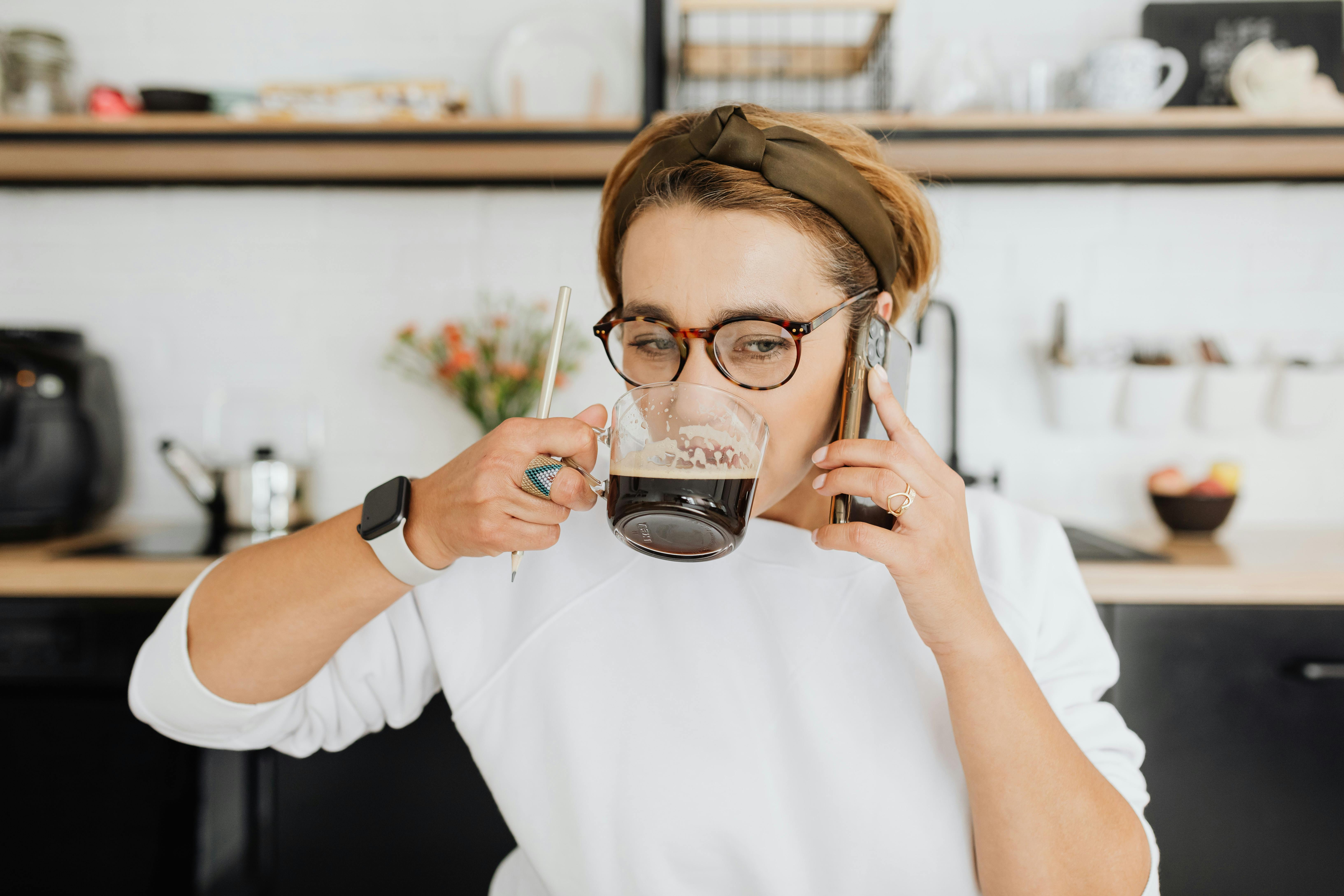 Woman multitasking, drinking coffee while on a phone call in a modern kitchen.