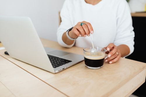 Free A woman stirs coffee at her desk while working on a laptop in a modern office setting. Stock Photo