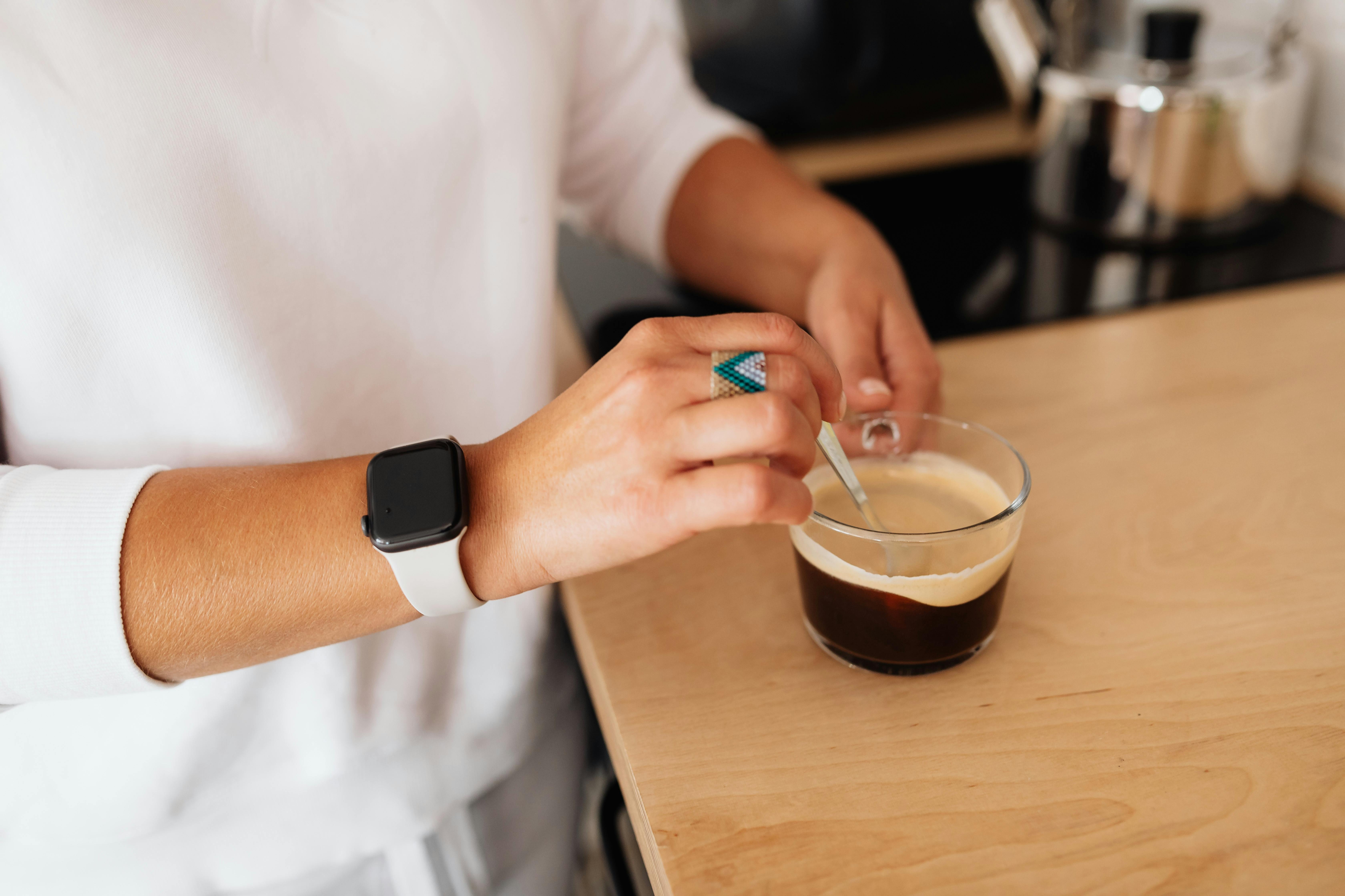 Close-up of a person stirring coffee in a glass cup, wearing smartwatch.