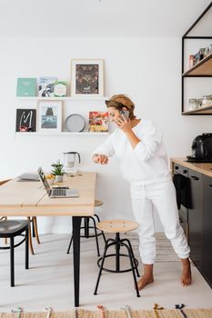 Adult woman multitasking at home with a laptop and phone call in a modern kitchen setting.