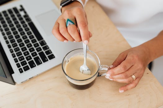 A person stirs coffee in a glass mug beside a laptop on a wooden table.
