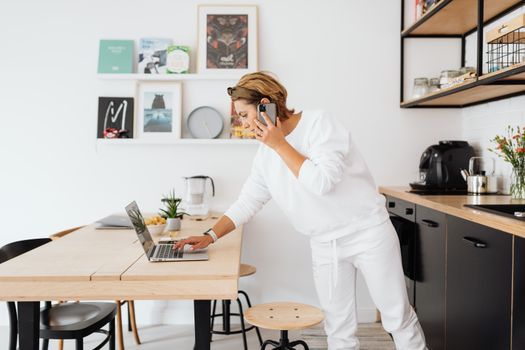 Woman using laptop and phone in stylish kitchen, blending work and home life.
