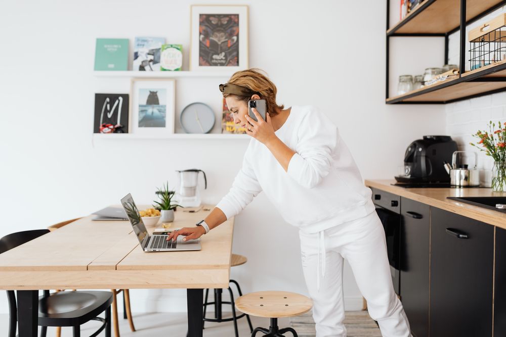 Woman using laptop and phone in stylish kitchen, blending work and home life.