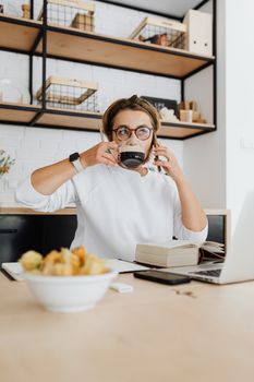Woman enjoying coffee while multitasking with a book and laptop in a modern kitchen setting.