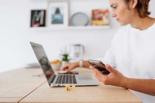 A woman in a white shirt using a laptop and smartphone at a wooden table.