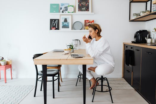 A woman multitasking with a smartphone and laptop in a modern kitchen, showcasing remote work lifestyle.