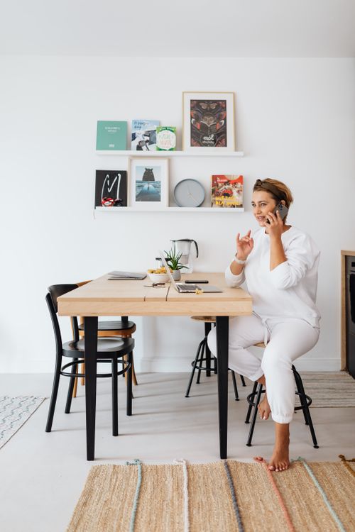 Free Woman working from home, talking on phone, sitting barefoot at a wooden table, interior design setting. Stock Photo