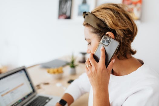 Focused woman multitasking with smartphone and laptop in a home office setting.