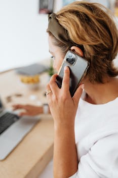 A woman uses a smartphone while working on a laptop at a desk, showcasing multitasking in a modern workspace.