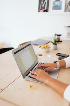 Close-up of woman typing on a laptop at a minimalist home desk with plants and office supplies.