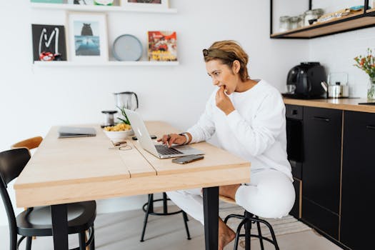 Casually dressed woman working on a laptop at home in a modern kitchen setting.