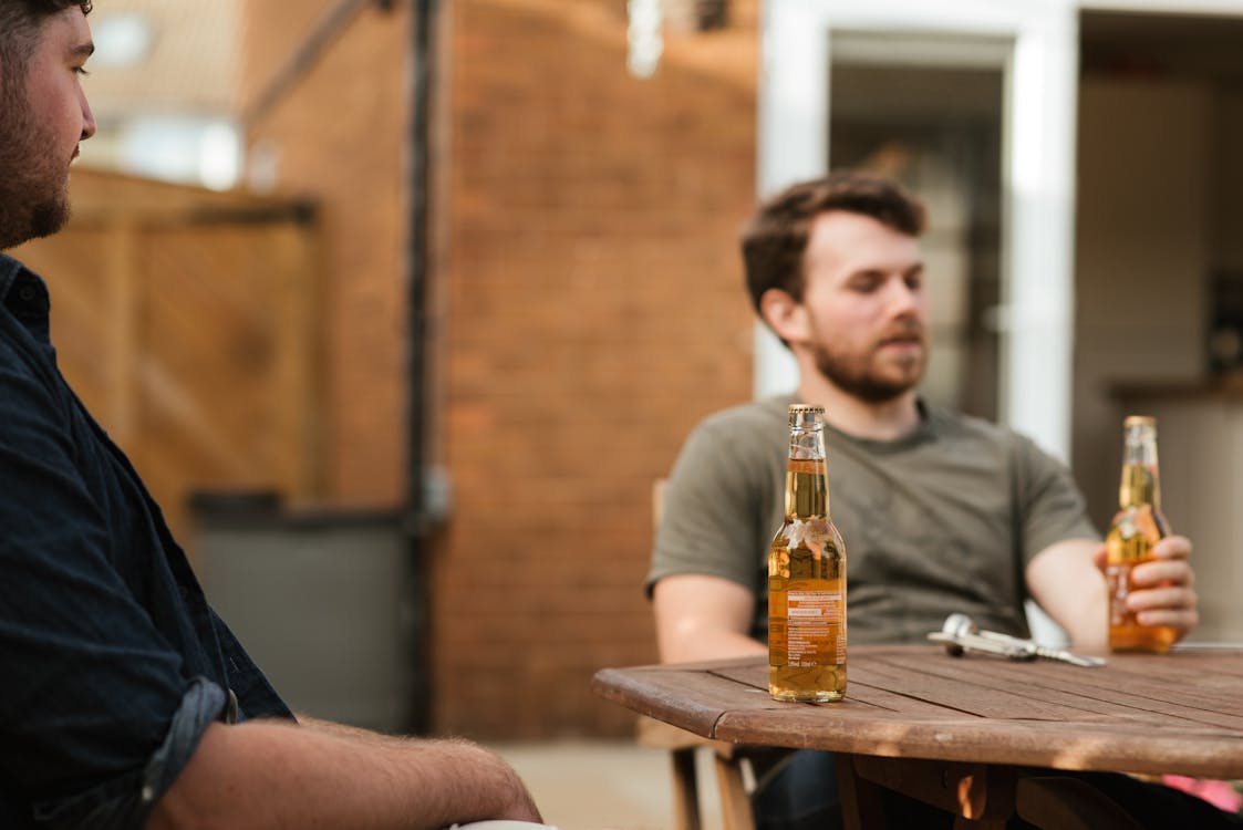 Adult bearded males friends sitting at table with bottles of beer while spending weekend together in yard
