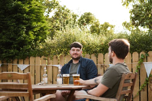 Adult mates chatting while resting with glass of beer at table in backyard in green countryside