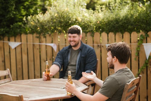 Smiling bearded adult men sitting with glass bottles of beer at wooden table in backyard in green countryside