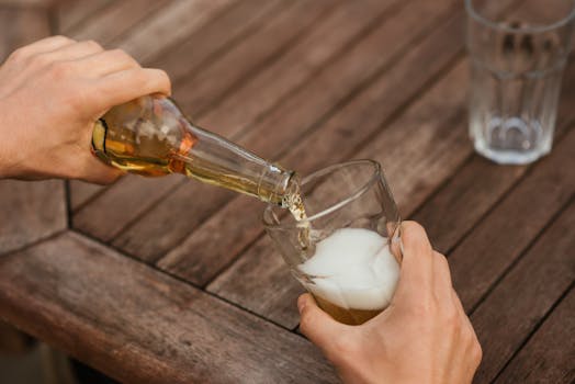 Person pouring beer from bottle into glass on an outdoor wooden table.