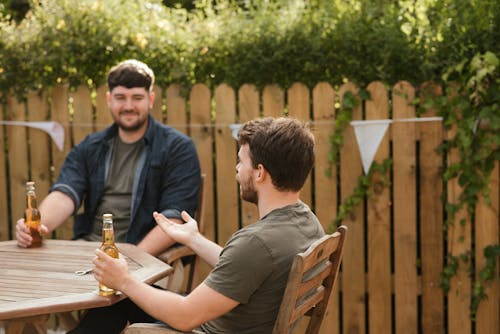 Positive bearded males wearing casual clothes sitting at table and drinking beer while resting in yard