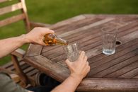 From above crop anonymous male sitting at wooden table and pouring cold beer into glass cup at weekend in yard