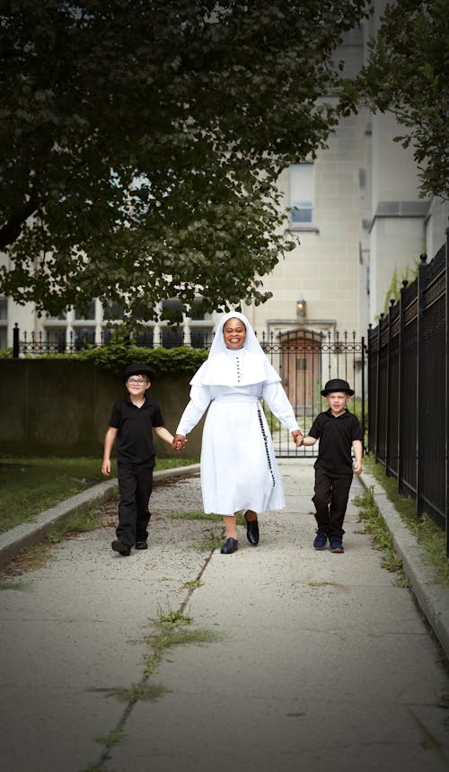 Free Full body of cheerful African American middle aged woman in traditional clothes holding hands with little boys in black outfits Stock Photo