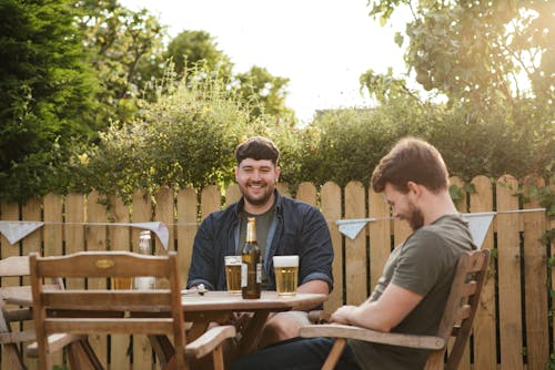 Laughing friends having beer while sitting in chairs at wooden table in backyard and having conversation