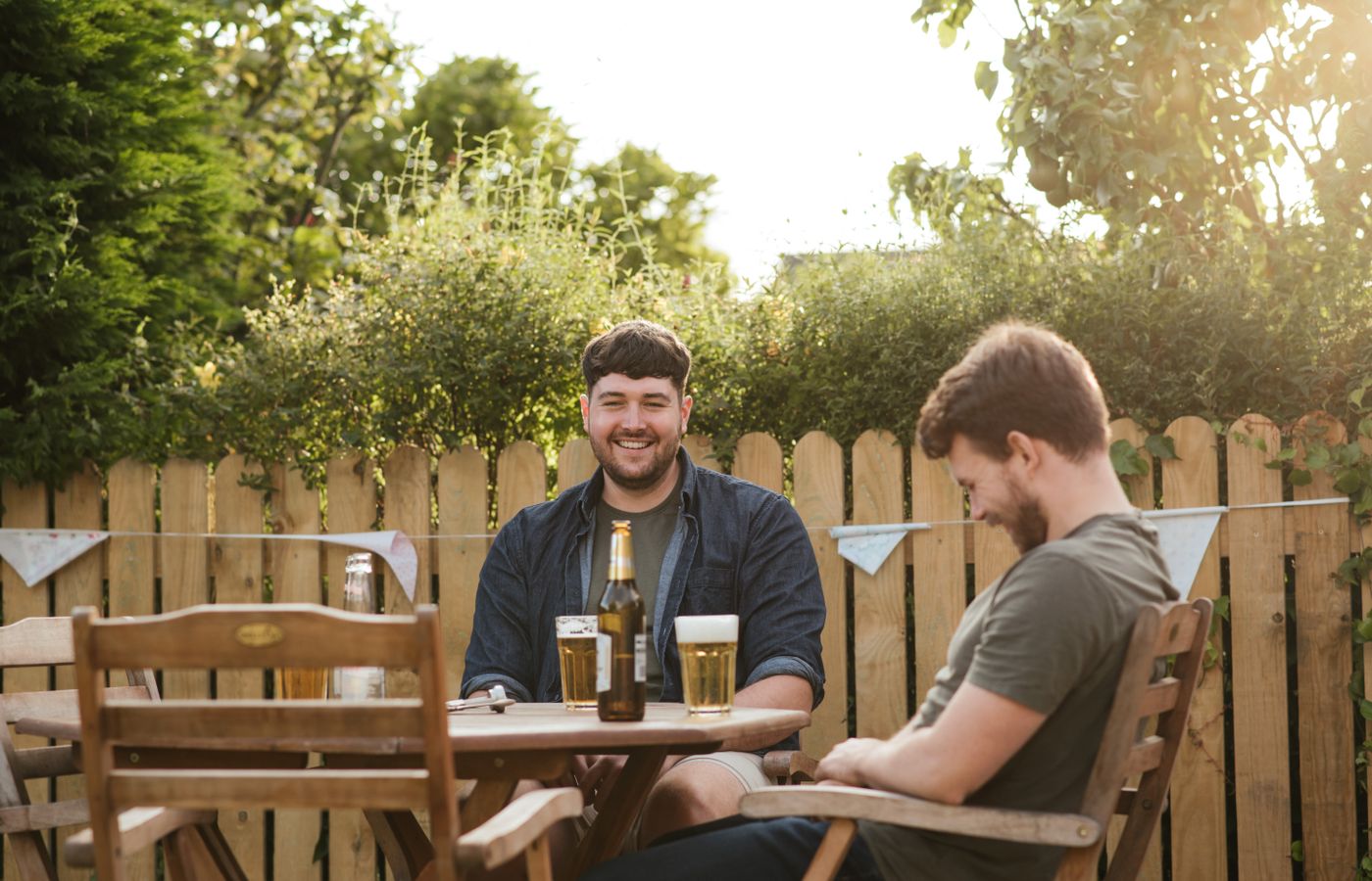 Two men relaxing with drinks in a sunny backyard setting, enjoying leisure time.