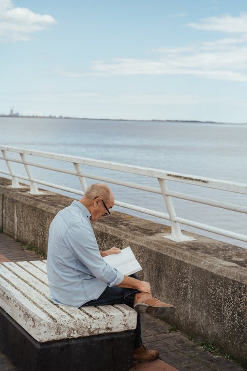 Free Side view of concentrated senior man in eyeglasses reading book while sitting on bench on waterfront Stock Photo