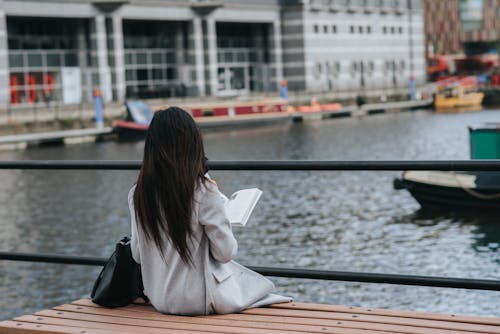 Free Back view of unrecognizable female in warm clothes reading book at spare time on waterfront Stock Photo