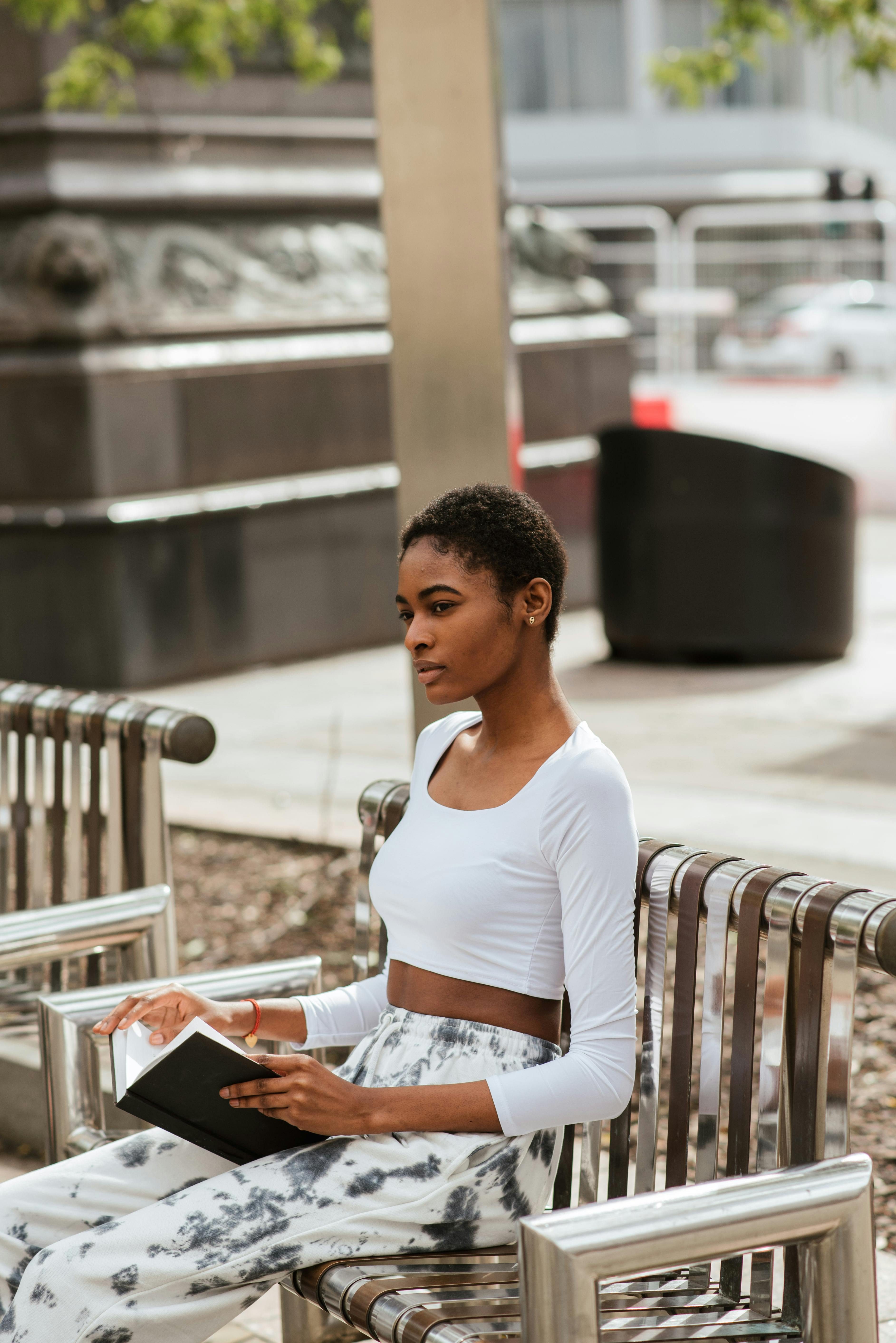 Thoughtful young African American female in white casual clothes with short dark hair sitting on bench in park and thinking over open book
