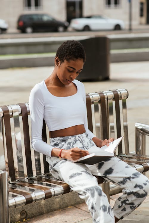 Young concentrated black female in casual apparel reading book while sitting on urban metal bench in sunlight