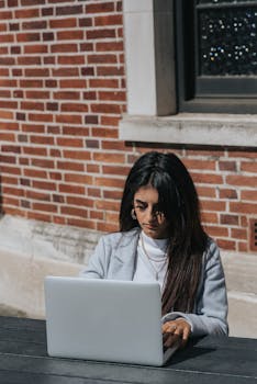 Young attentive ethnic female distance worker typing on portable computer at table near brick building