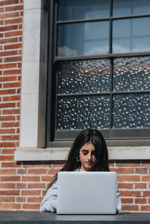 Young ethnic female executive watching netbook at table near brick building on summer day